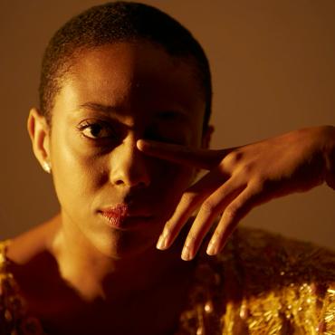 A brown skin Black woman with a buzz cut stares at the camera with her right eye wide open and left eye closed by her left index finger. She is wearing a shimmering orange top, gold lightning bolt earrings, and pink lipstick.